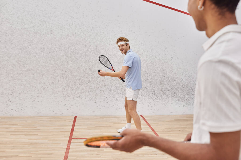 enfoque en el hombre feliz en ropa deportiva jugando con un amigo afroamericano dentro de la cancha de squash - Foto, Imagen