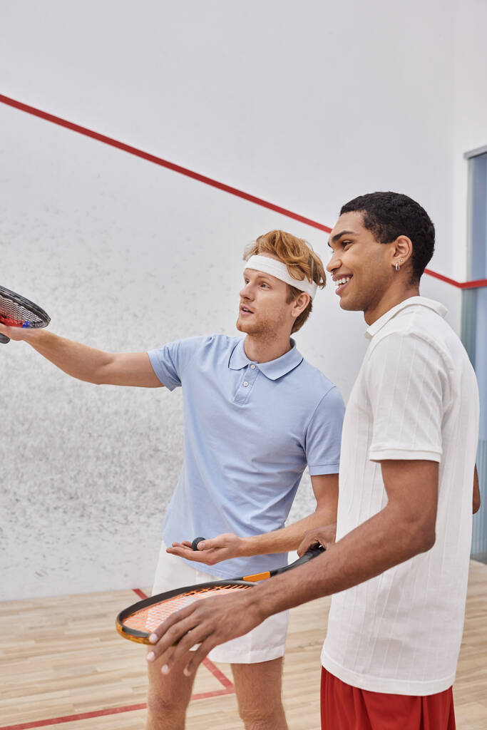 felices jugadores interracial en ropa deportiva sonriendo y mirando hacia el interior de la pista de squash, raquetas - Foto, Imagen