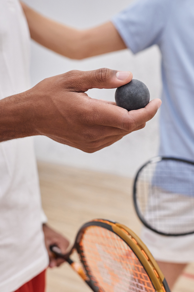 tiro recortado de hombre afroamericano sosteniendo pelota de squash cerca de amigo dentro de la cancha, vertical - Foto, Imagen