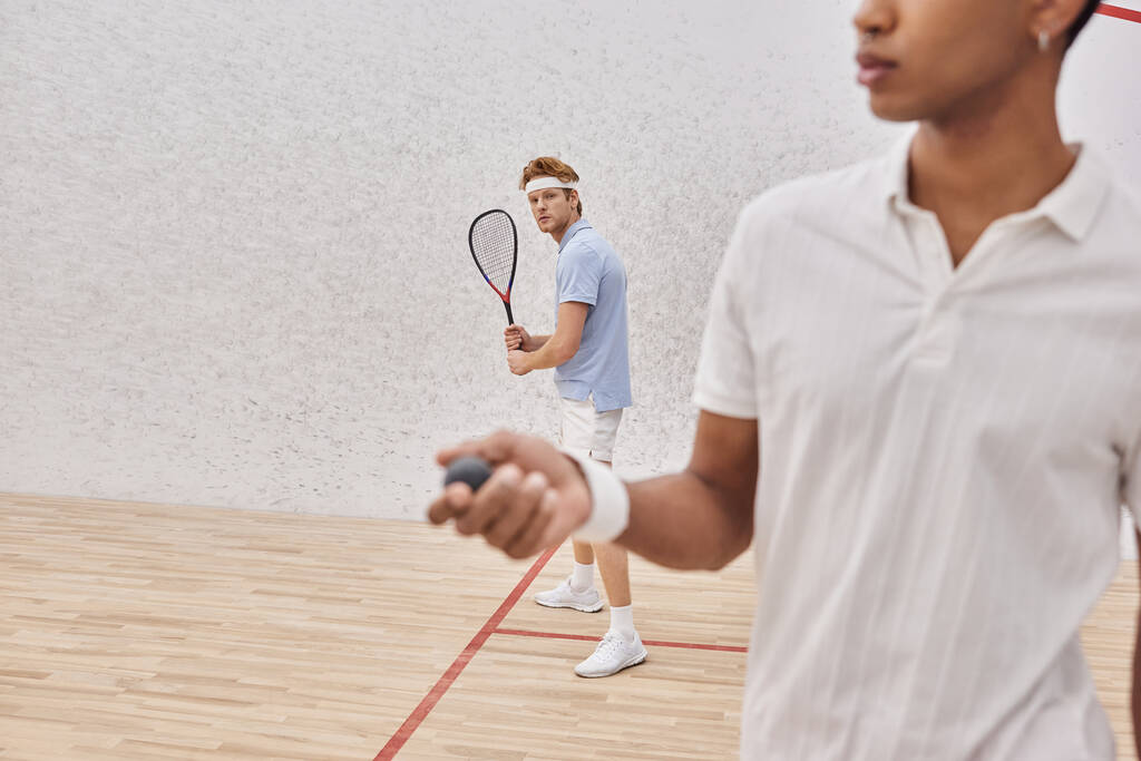pelirroja sosteniendo raqueta cerca de hombre afroamericano con pelota de squash jugando en corte - Foto, imagen