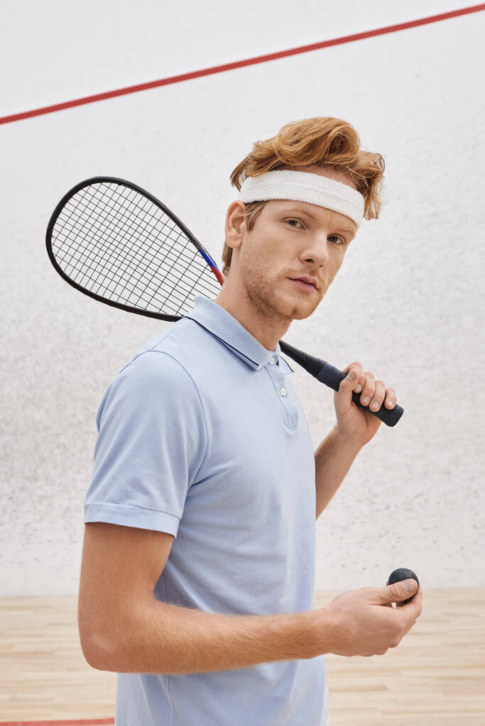pelirrojo en ropa deportiva sosteniendo pelota de squash y raqueta mientras está de pie dentro de la cancha, vertical - Foto, imagen