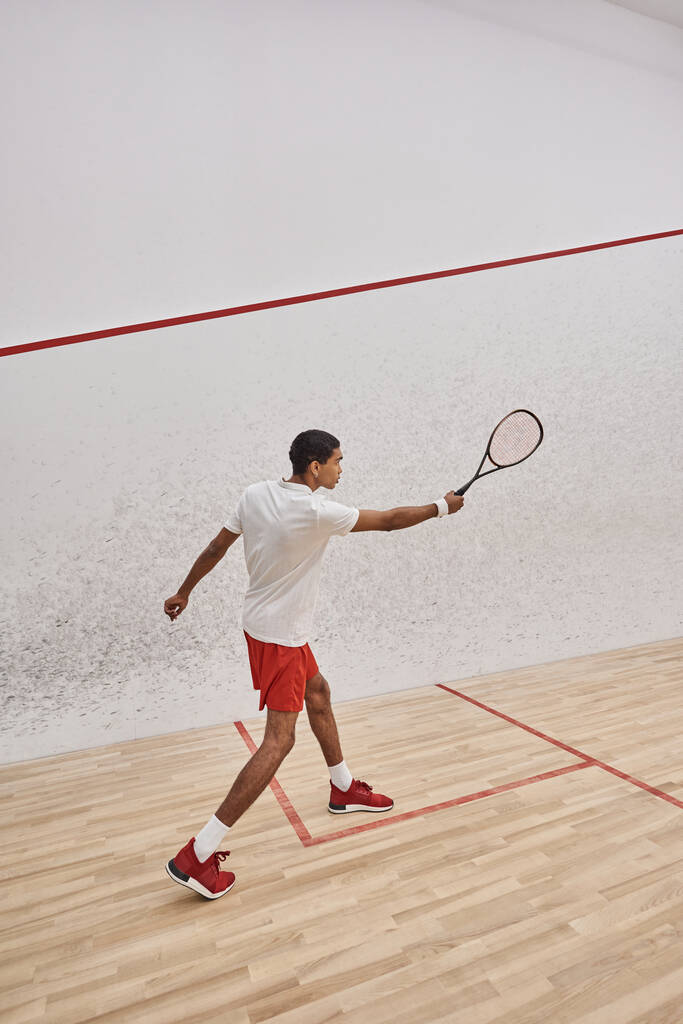 athletischer afrikanisch-amerikanischer Sportler hält Squash-Schläger in der Hand und springt, während er ein Spiel vor Gericht spielt - Foto, Bild