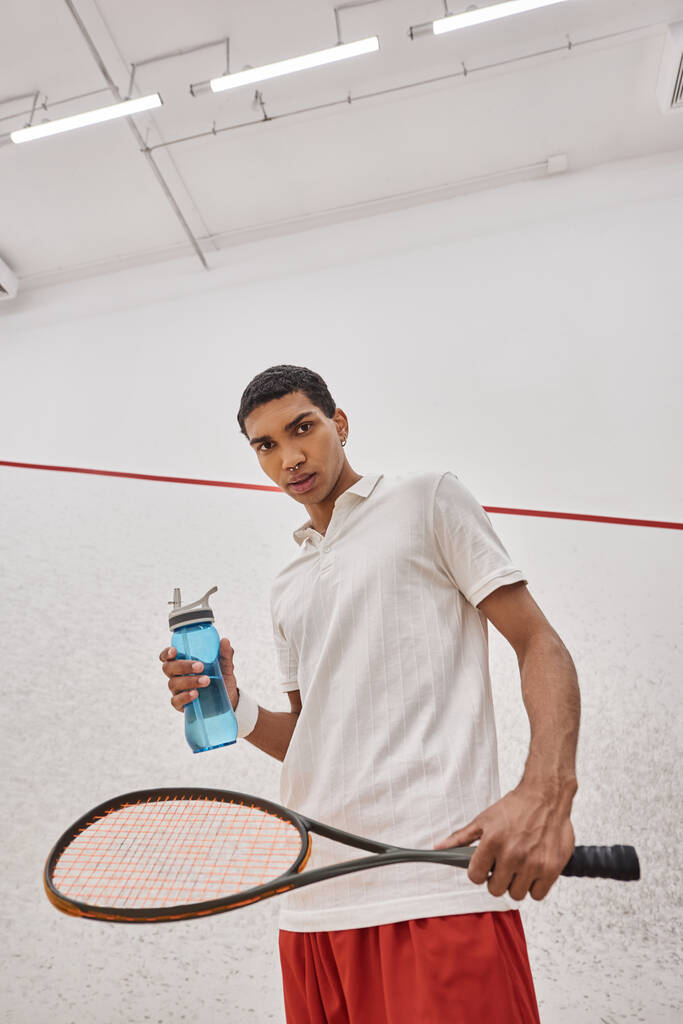 νεαρός Αφροαμερικανός αθλητής κρατώντας ένα μπουκάλι νερό και σκουός racquet μετά το παιχνίδι - Φωτογραφία, εικόνα