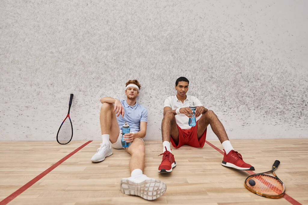 two tired interracial friends with squash racquets and bottle with water sitting on floor in court - Photo, Image