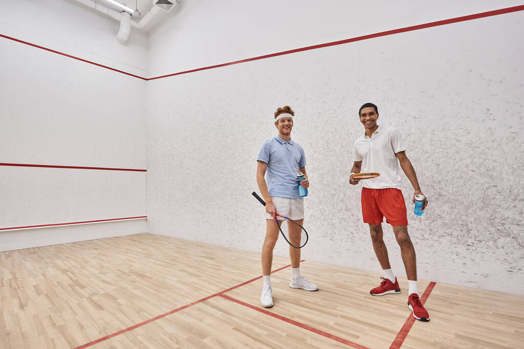 allegri sportivi interrazziale con racchette da squash e bottiglie con acqua in piedi all'interno della corte - Foto, immagini