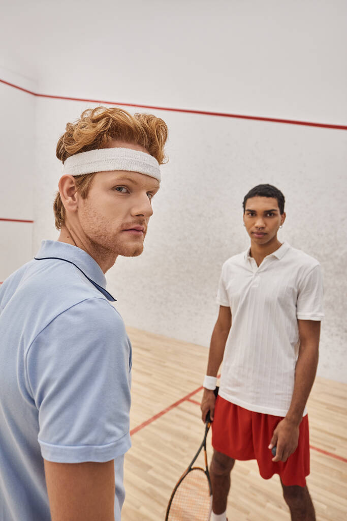 skoncentrowany międzyrasowy mężczyzna przyjaciele w odzież sportowa stojąc razem z squash rakiety w sądzie - Zdjęcie, obraz