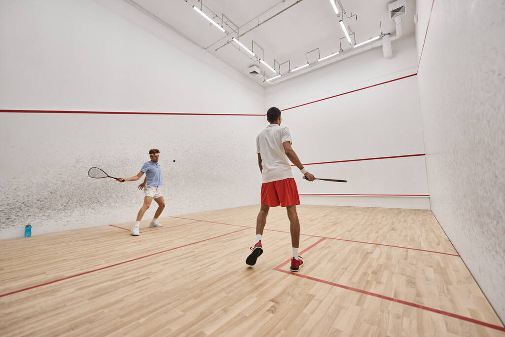 hommes interraciaux et sportifs en vêtements de sport jouant au squash ensemble à l'intérieur du court, motivation - Photo, image
