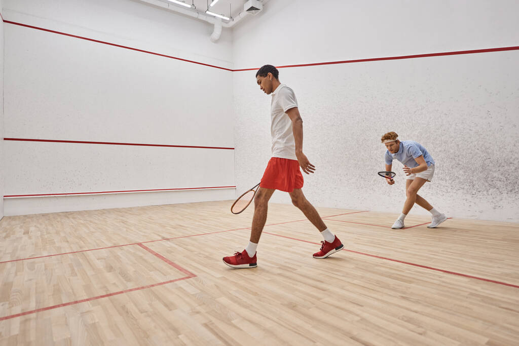 aktivní a různorodí muži ve sportovním oblečení hrát squash uvnitř soudu, výzva a motivace - Fotografie, Obrázek