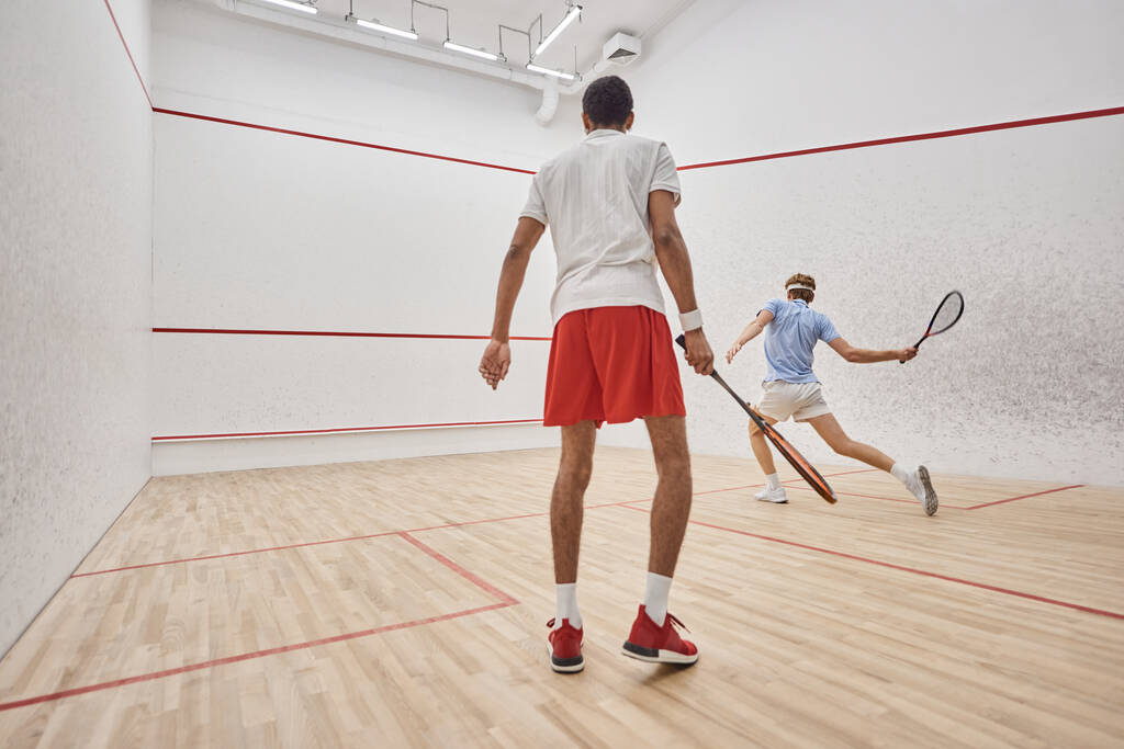 hombres activos interracial en ropa deportiva jugando squash dentro de la cancha, desafío y motivación - Foto, Imagen