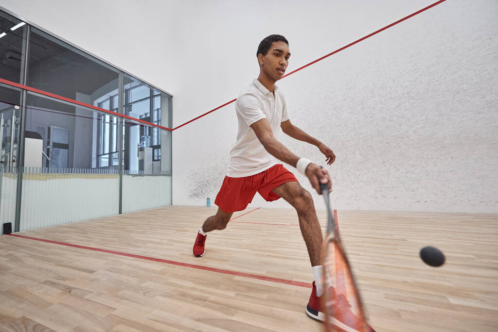 deportista afroamericano en pantalones cortos deportivos rojos sosteniendo raqueta mientras juega squash, tiro de movimiento - Foto, Imagen