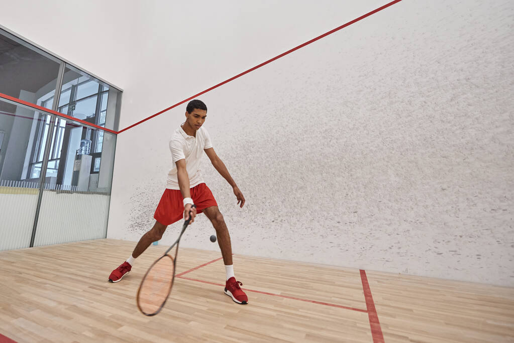 jonge Afro-Amerikaanse man in rode sportieve shorts houden racket tijdens het spelen squash, motion shot - Foto, afbeelding