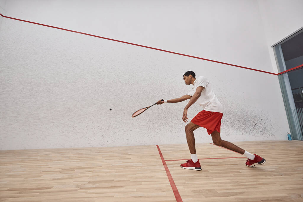 aktywny afrykański sportowiec w czerwonych spodenkach trzymający rakietę podczas gry w squasha w sądzie - Zdjęcie, obraz