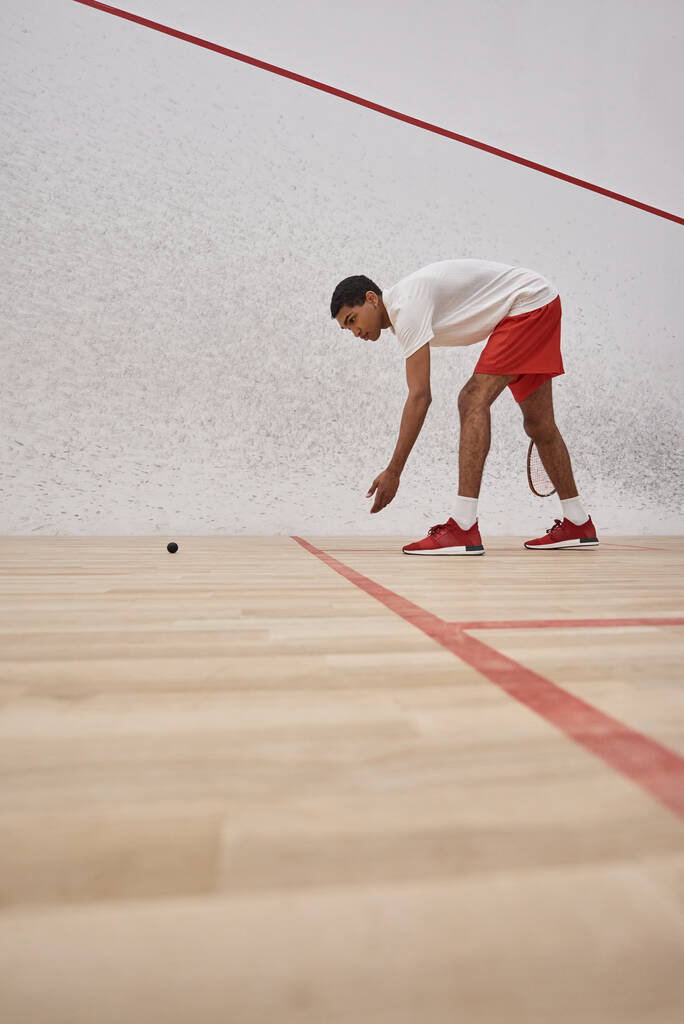 homem americano africano em shorts esportivos vermelhos segurando raquete e pegando bola de squash dentro da quadra - Foto, Imagem