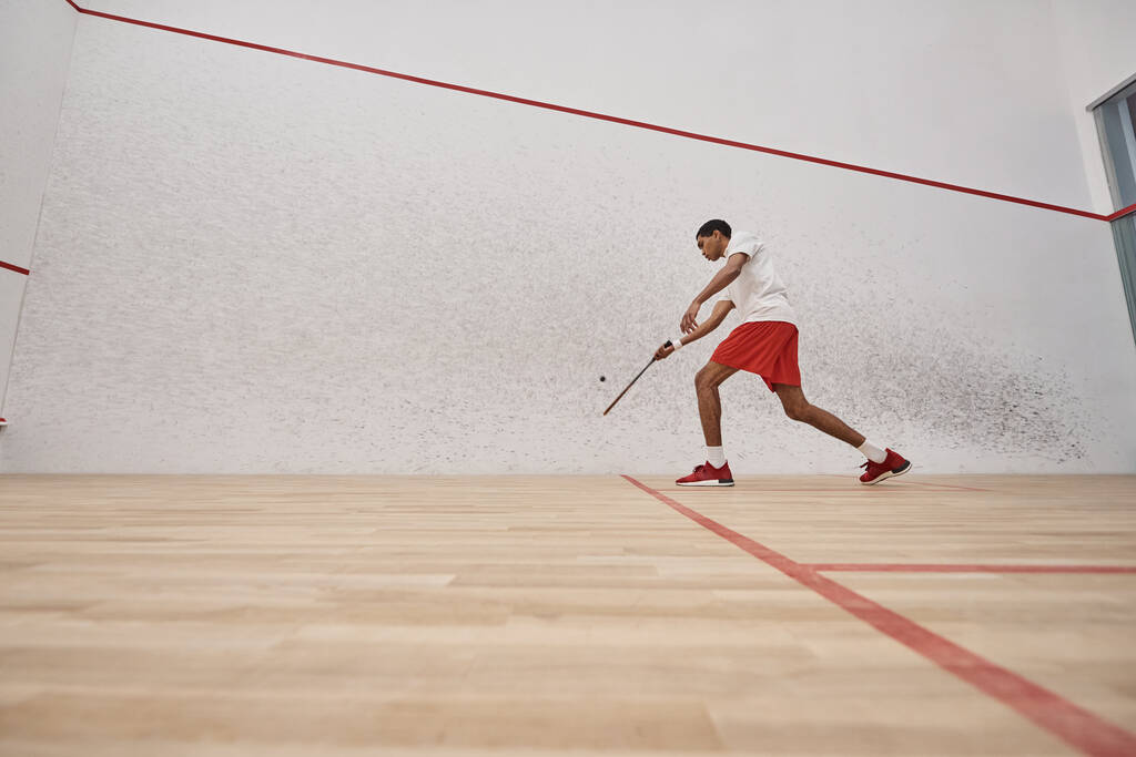 afro-américain jeune homme en short rouge tenant raquette tout en jouant à la courge à l'intérieur de la cour - Photo, image