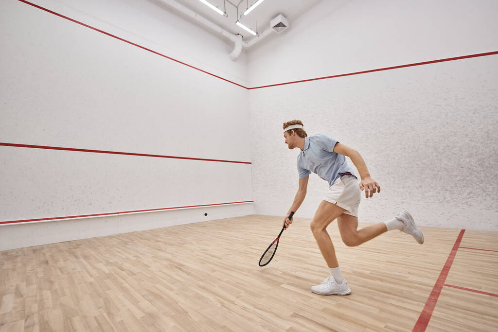 urheilullinen ja punapää urheilija maila pelaa squash sisällä tuomioistuin, liike laukaus - Valokuva, kuva