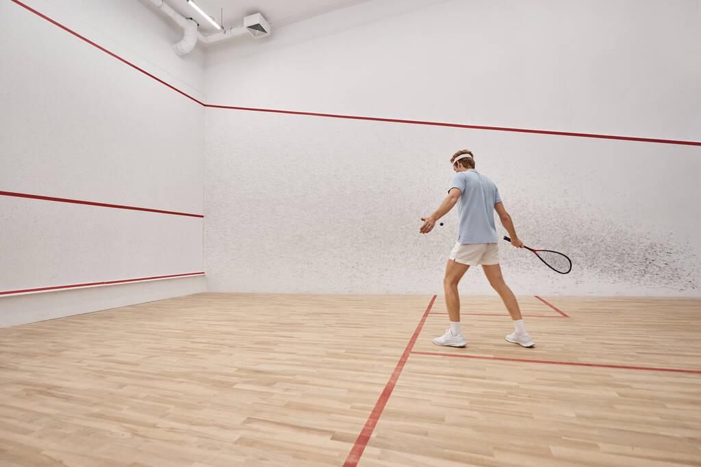 uomo sportivo e rosso che tiene la racchetta mentre gioca a squash all'interno della corte, motion shot - Foto, immagini