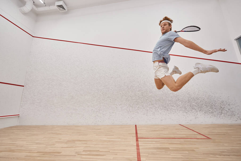 tiro en movimiento, deportista enérgico sosteniendo raqueta y saltando mientras juega squash dentro de la cancha - Foto, Imagen