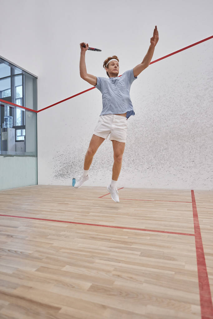 pohybová fotografie, legrační sportovec drží raketu a skákání při hraní squash uvnitř soudu - Fotografie, Obrázek