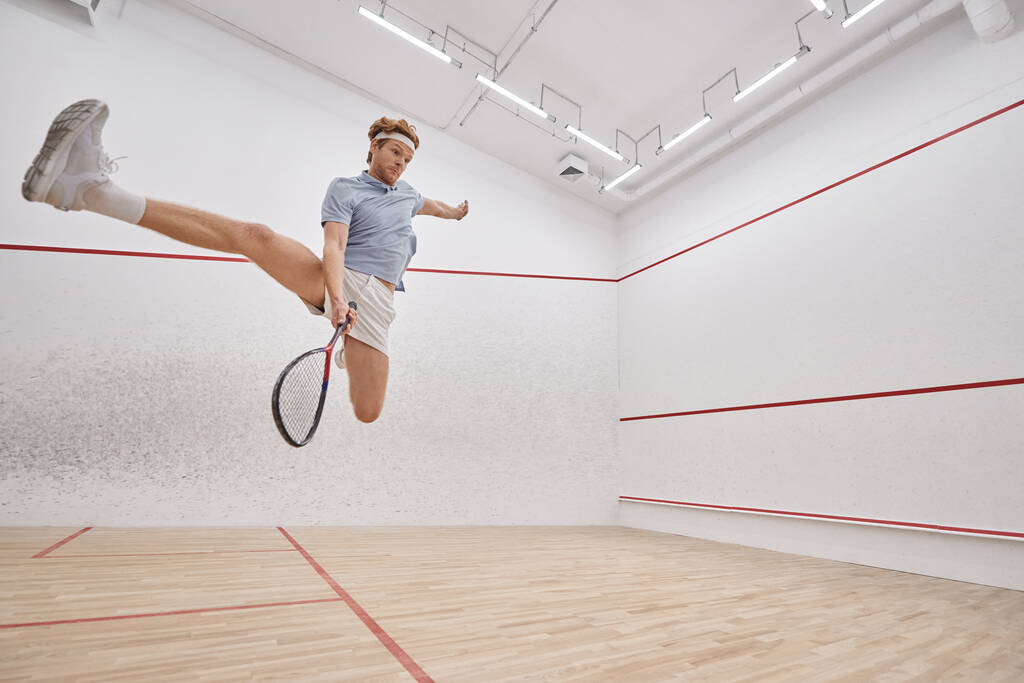 disparo de movimiento, jugador enérgico sosteniendo raqueta mientras salta y juega squash dentro de la cancha - Foto, imagen
