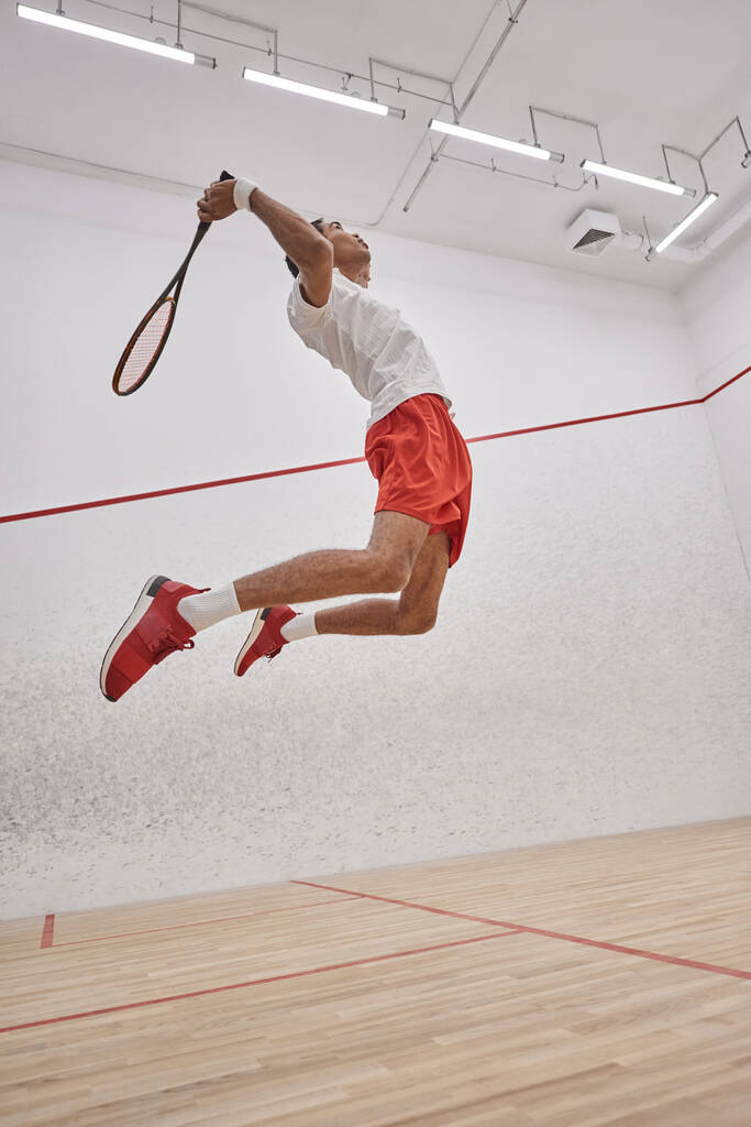 фотографія руху, активний афроамериканський спортсмен, який тримає ракетку під час стрибків і гри в сквош - Фото, зображення