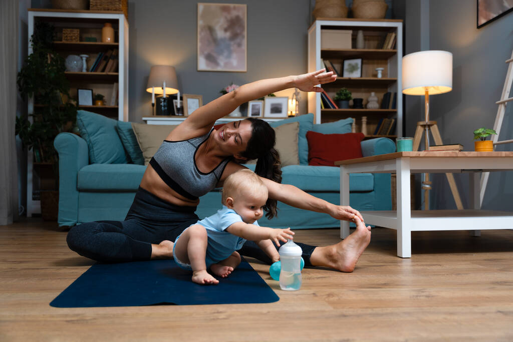 Die junge glückliche, erfolgreiche, alleinerziehende Mutter praktiziert zu Hause Yoga auf einer Yogamatte, während ihr Baby achtlos auf dem Boden sitzt und mit einer Wasserflasche spielt. Mama dehnt Muskeln - Foto, Bild