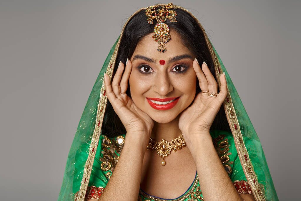 veselá mladá indická žena s tečkami a zeleným závojem usmívající se na kameru s rukama u obličeje - Fotografie, Obrázek