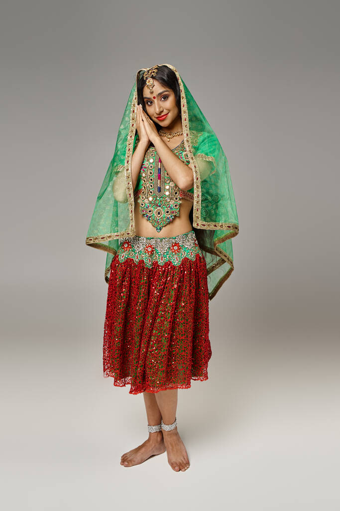 iloinen intialainen nainen punaisessa hameessa ja vihreässä kolissa poseeraa harmaalla taustalla ja katselee kameraa - Valokuva, kuva