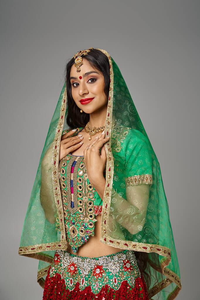 tiro vertical de mujer india joven en ropa tradicional y velo verde posando sobre fondo gris - Foto, Imagen