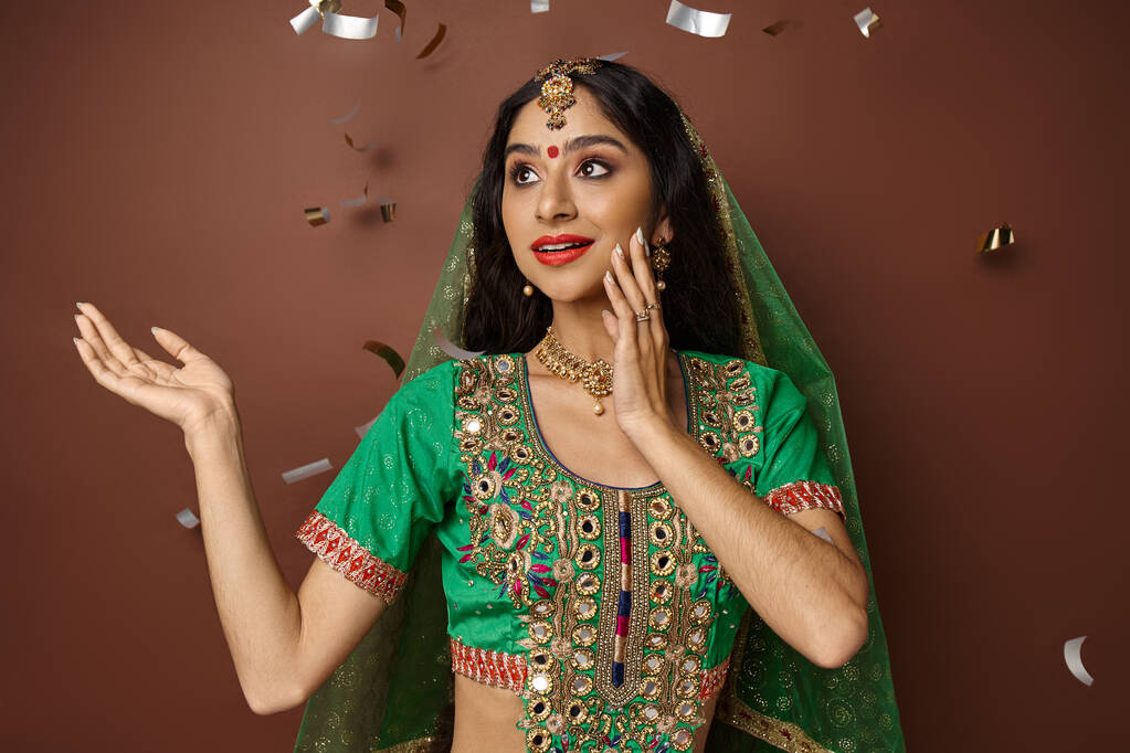 fröhliche hübsche Indianerin in traditioneller Kleidung posiert vor braunem Hintergrund unter Konfettiregen - Foto, Bild