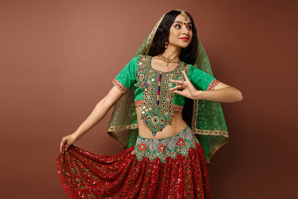 привлекательная индийская женщина с бинди в традиционной одежде жестикулируя во время танцев на коричневом фоне - Фото, изображение
