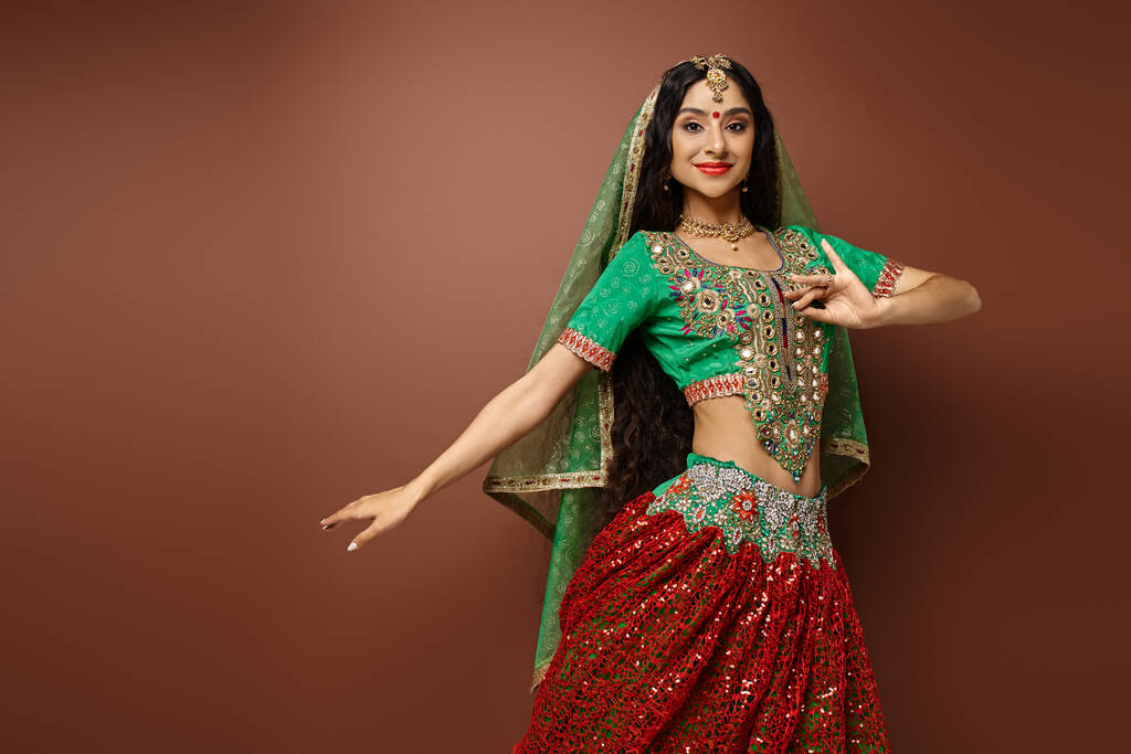 radosna młoda Hinduska kobieta w zielonym choli z bindi dot gestykulując podczas tańca patrząc w kamerę - Zdjęcie, obraz