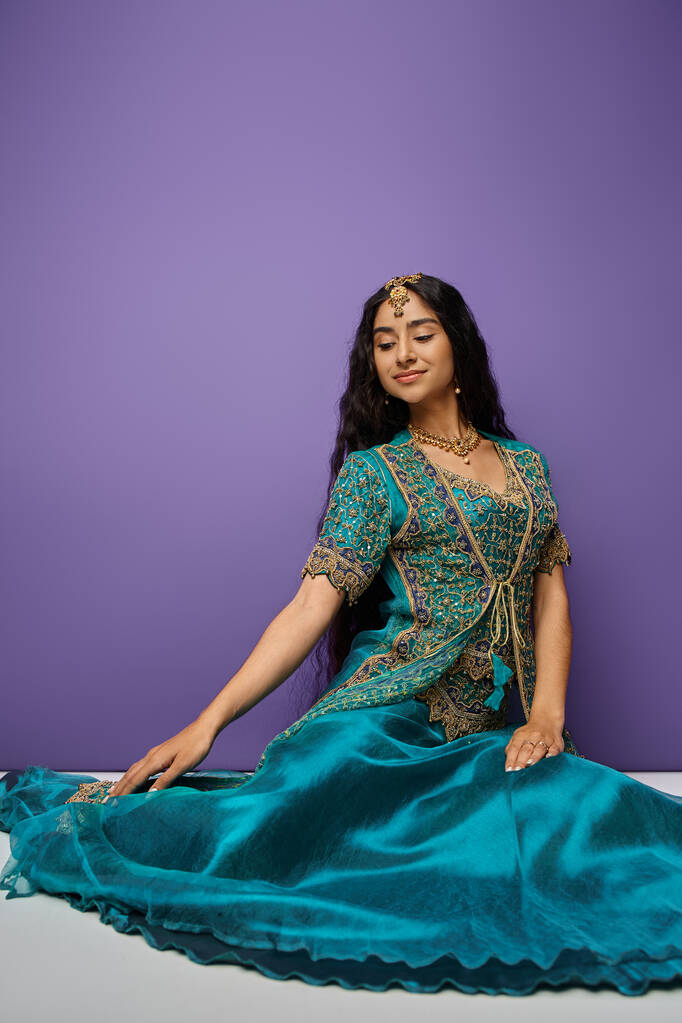 pionowy strzał atrakcyjnej hinduskiej kobiety siedzącej na podłodze w niebieskim tradycyjnym sari i odwracając wzrok - Zdjęcie, obraz