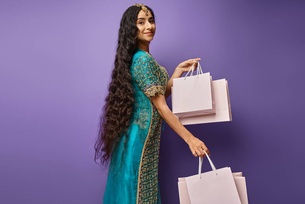 Oldukça neşeli, uzun saçlı, mavi elbiseli, mor arka planda alışveriş torbalarıyla poz veren Hintli bir kadın. - Fotoğraf, Görsel