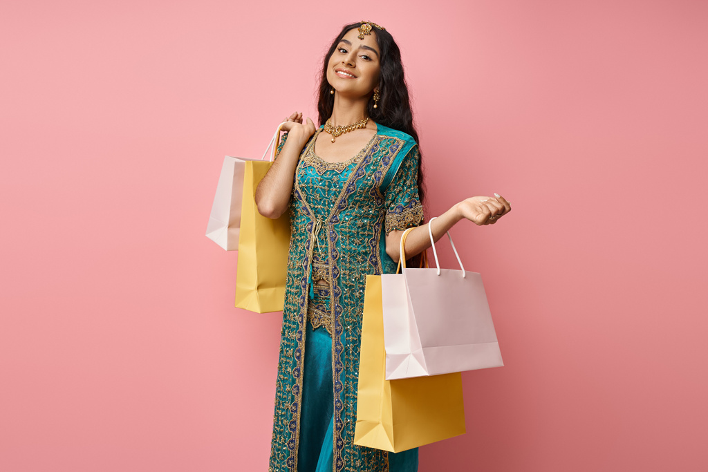 Ulusal kostümlü neşeli Hintli kadın pembe arka planda alışveriş torbalarıyla poz veriyor. - Fotoğraf, Görsel