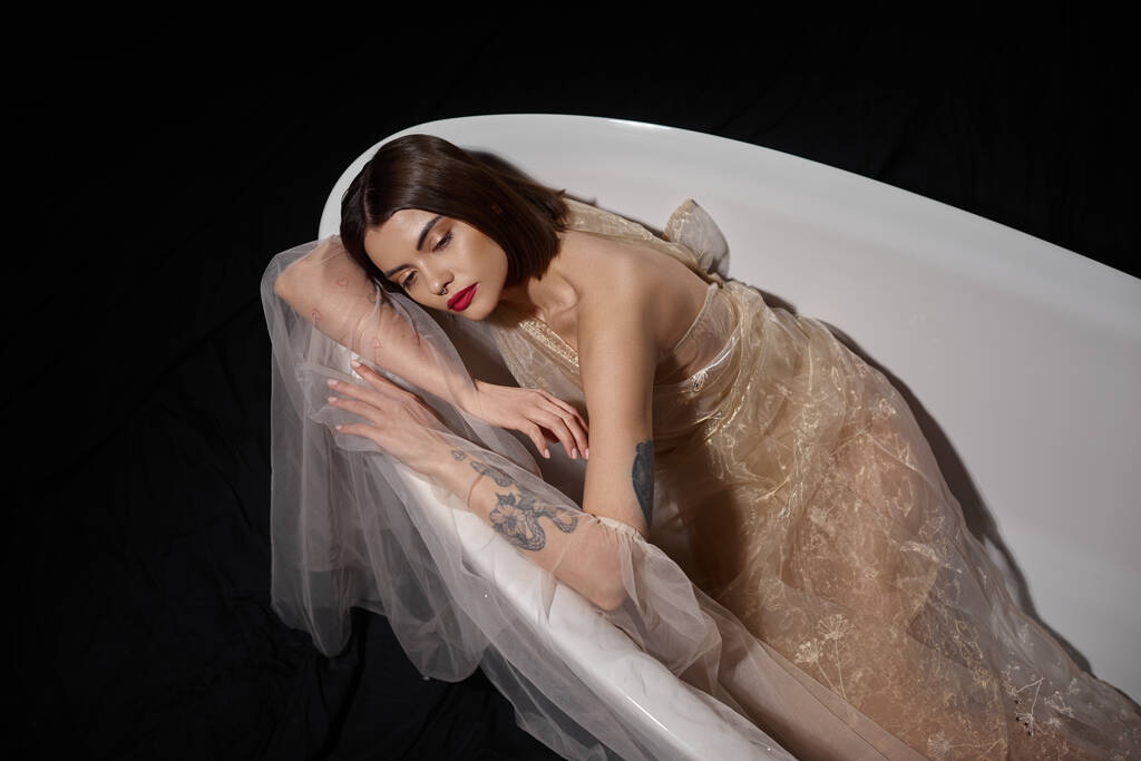 high angle view of graceful young woman in transparent dress while lying on bathtub on black - Photo, Image