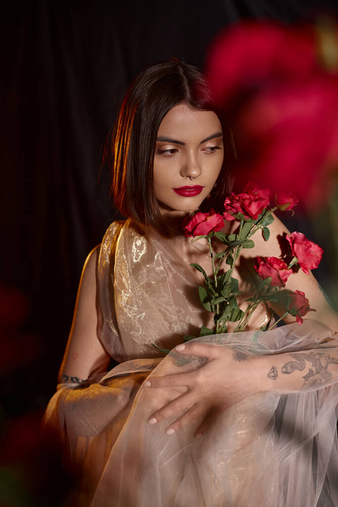 чувственная молодая женщина в романтическом прозрачном платье держа цветущие красные розы на черном фоне - Фото, изображение