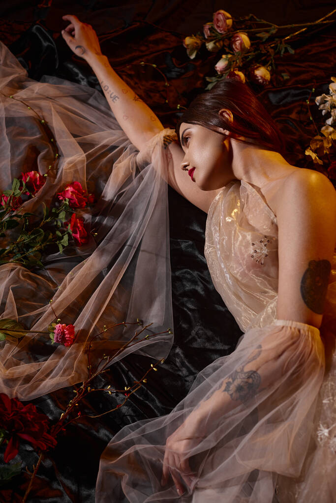 krásná a tetovaná mladá žena v romantických průhledných šatech ležící mezi kvetoucími květinami - Fotografie, Obrázek