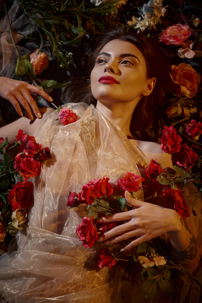 weibliche Anmut, tätowierte Frau in romantischem transparentem Kleid, die zwischen wunderschönen Blumen liegt - Foto, Bild