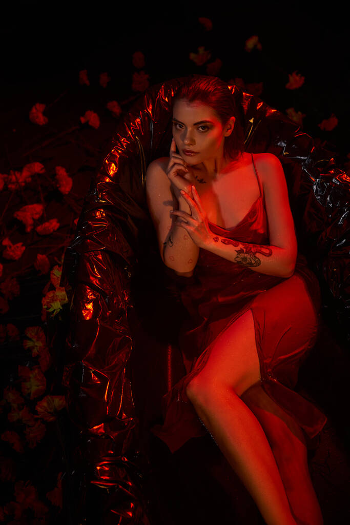 pohled shora, mladá smyslná žena v červené kombiné šaty ležící v černé vaně mezi krásné květiny - Fotografie, Obrázek
