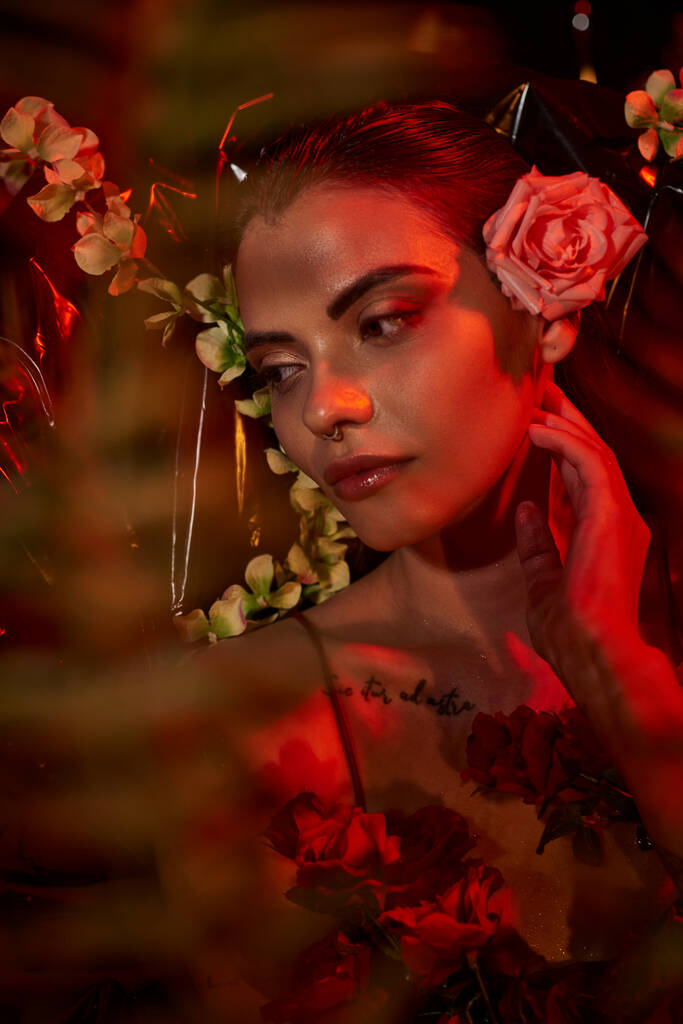 Farklı çiçeklerin arasında duran piercing ve dövmeli rüya gibi genç bir kadının yüzünde kırmızı ışık. - Fotoğraf, Görsel