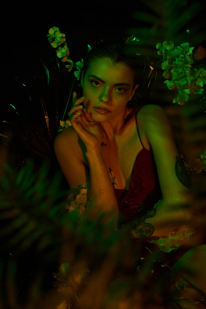 luz verde, mujer joven atractiva con el pelo mojado mirando a la cámara y posando entre flores - Foto, imagen