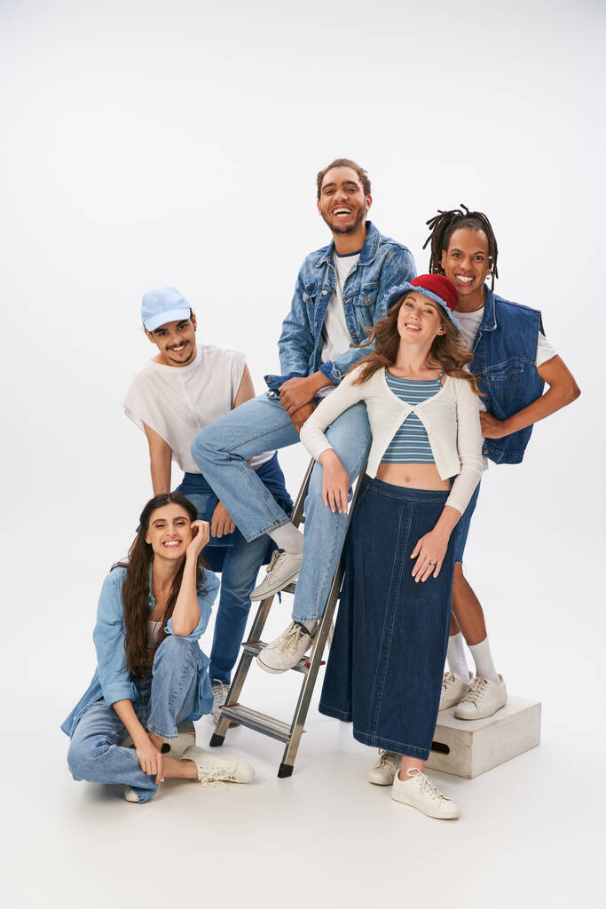 fröhliche multikulturelle Freunde in trendigen Klamotten posieren mit Leiter und blicken in die Kamera auf grau - Foto, Bild
