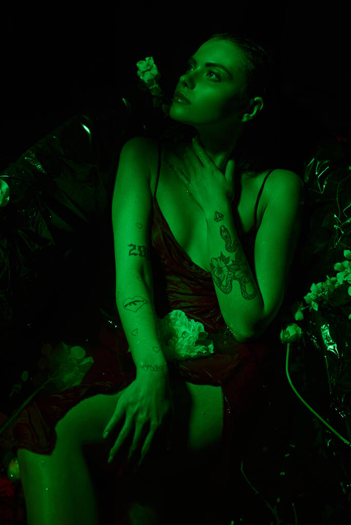 πράσινο φως, ονειρική και νεαρή γυναίκα με βρεγμένα μαλλιά κοιτάζει μακριά και ποζάροντας ανάμεσα σε λουλούδια στην μπανιέρα - Φωτογραφία, εικόνα