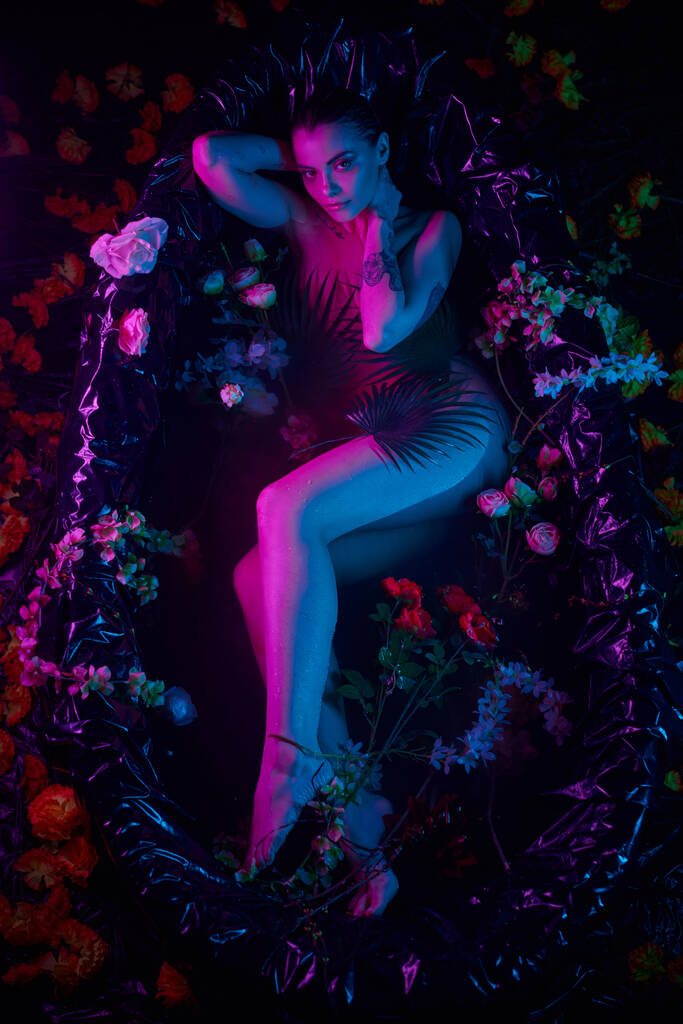 vrouwelijke schoonheid, jonge vrouw liggend tussen palmbladeren en bloemen in bad, blauw en paars licht - Foto, afbeelding