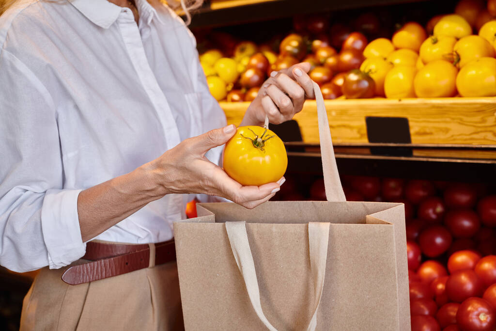 vista ritagliata di donna matura mettendo pomodoro giallo nella borsa della spesa mentre al negozio di alimentari - Foto, immagini