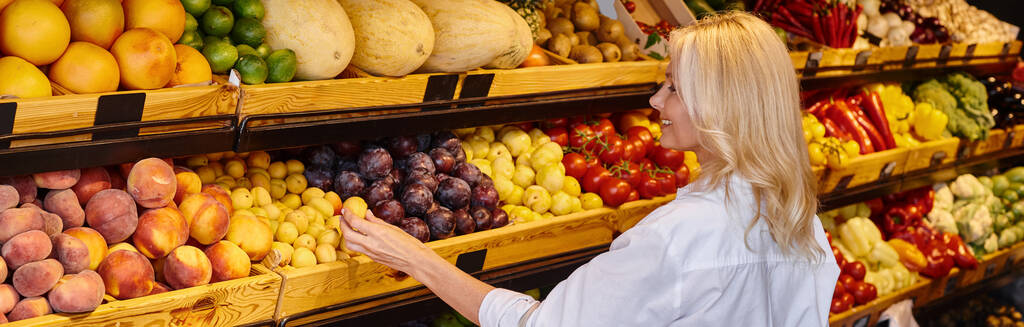 wesoła dojrzała kobieta w luźnych ubraniach z torbą na zakupy wybierając owoce w sklepie spożywczym, baner - Zdjęcie, obraz