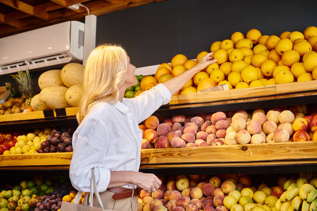érett szőke örömteli nő a mindennapi ruházat bevásárló táska szedés narancs élelmiszer boltban - Fotó, kép