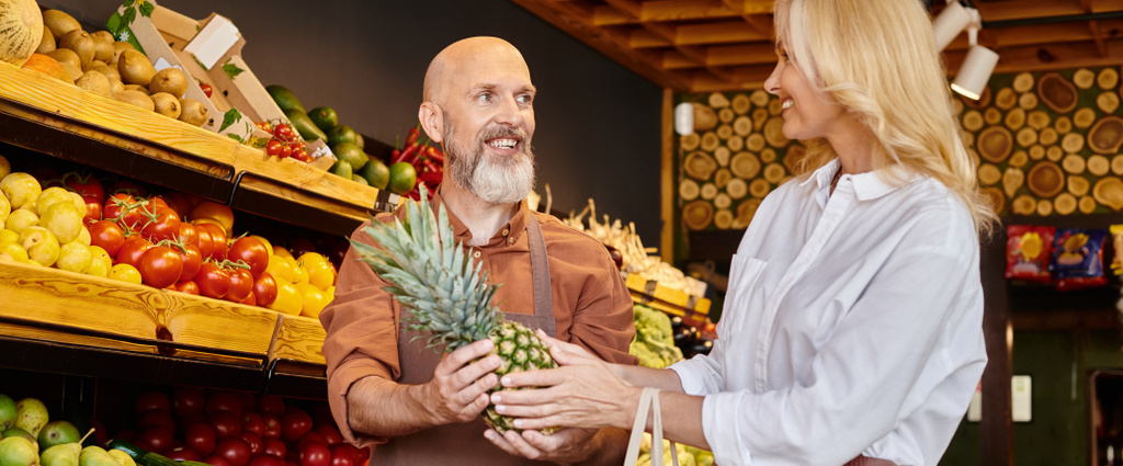radosny brodaty sprzedawca daje świeży ananas dla jego dojrzałej klientki w sklepie spożywczym, baner - Zdjęcie, obraz