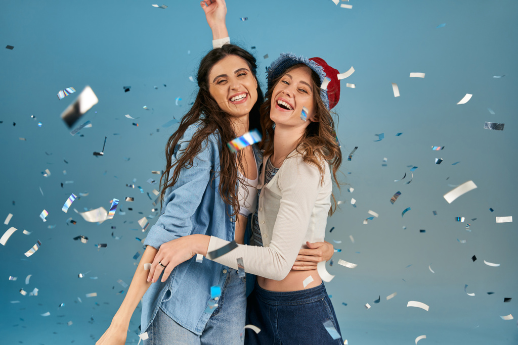 zorgeloze en trendy vriendinnen omarmen onder sprankelende confetti regen op blauwe achtergrond, feest tijd - Foto, afbeelding