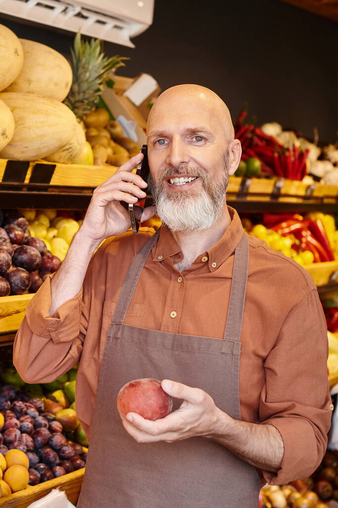 vertical shot of gray bearded jolly seller talking by phone and smiling at camera with peach in hand - Photo, Image